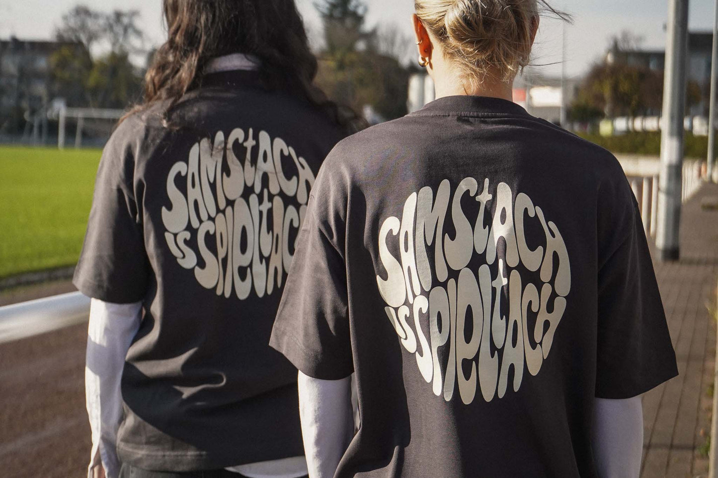 Zwei junge Frauen stehen am Spielfeldrand eines Fußballplatzes und tragen das "samstach is spieltach" Shirt in Stone Grey, so dass man den Backprint lesen kann.