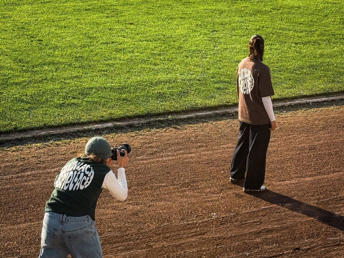 Das Bild zeigt Gründerin Meret, die ein Model mit dem "sonntach is spieltach" Shirt in Chocolate auf dem Fußballplatz fotografiert.