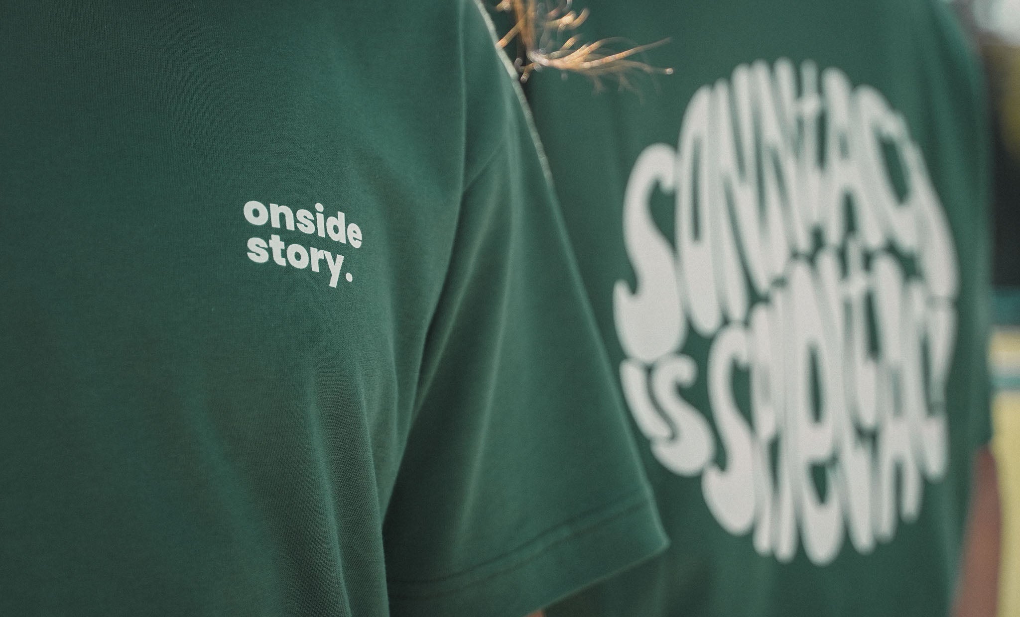 Detailansicht der nachhaltig und fair produzierten Shirts in Bottlegreen mit Backprint "sonntach is spieltach" und Logo auf der Brust für die Fußball-Community.