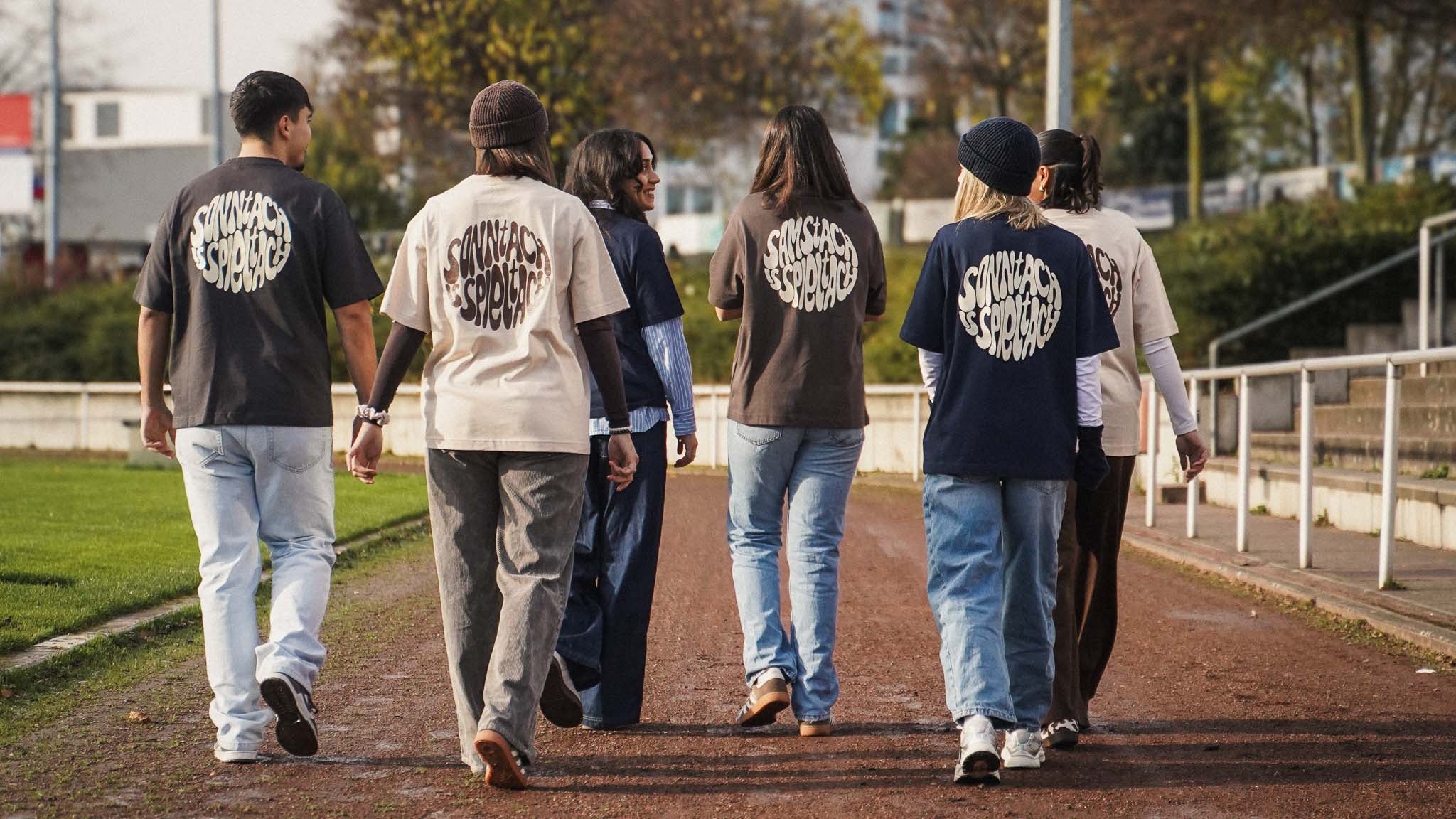 Eine Gruppe junger Fußballfans geht an der Seitenlinie eines Fußballplatzes entlang und trägt alle Varianten der neuen Shirts aus Chapter Two.