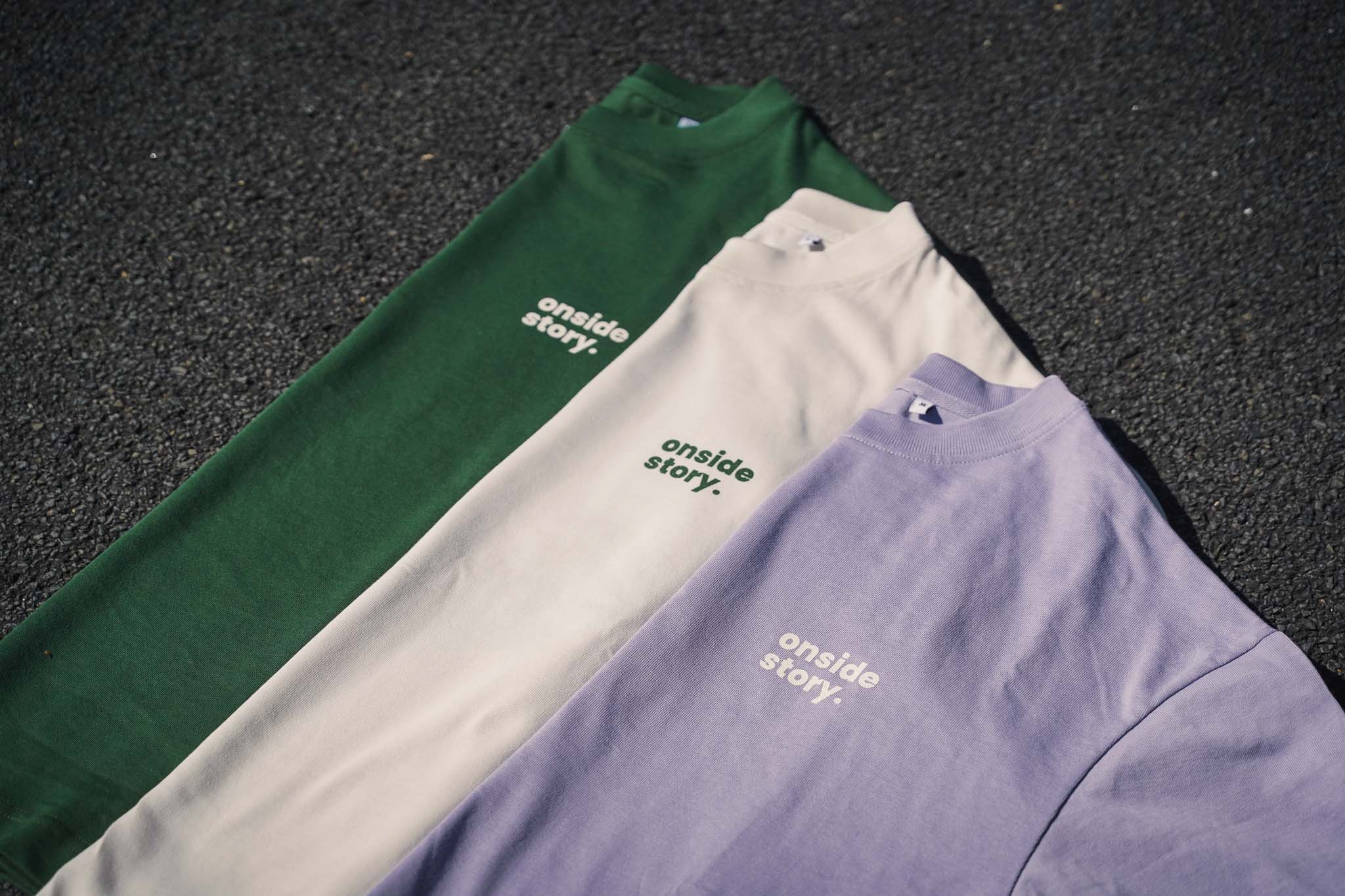 Drei Shirts in den Farben Bottlegreen, Cream und Lavender liegen gefaltet auf dem Asphalt. Man sieht das prägnante Logo der Streetwear Marke onside story.