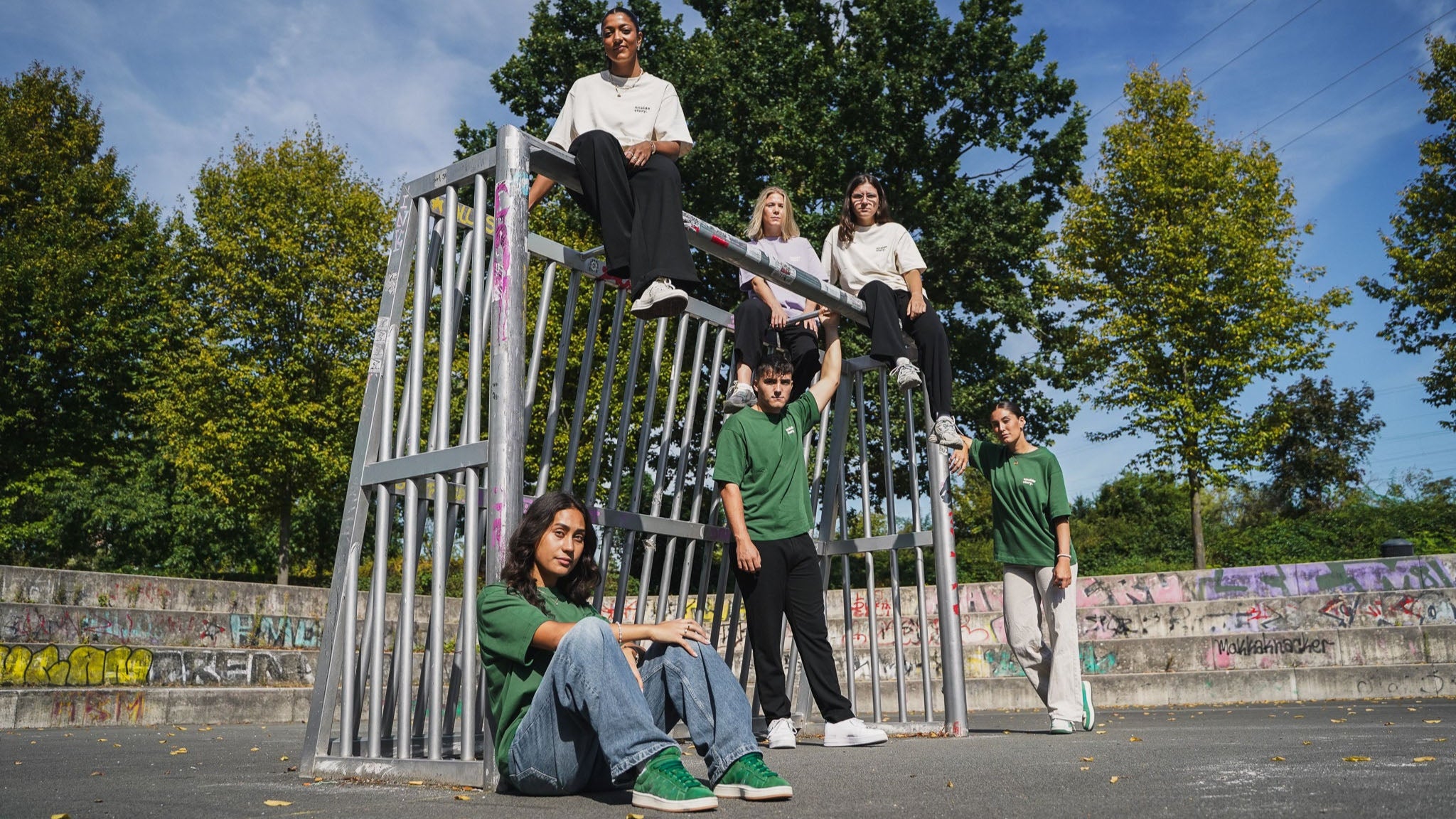 Gruppe junger Menschen trägt onside story Streetwear T-Shirts in Bottlegreen, Cream und Lavender, aufgenommen auf einem Bolzplatz mit Graffiti und Tor.