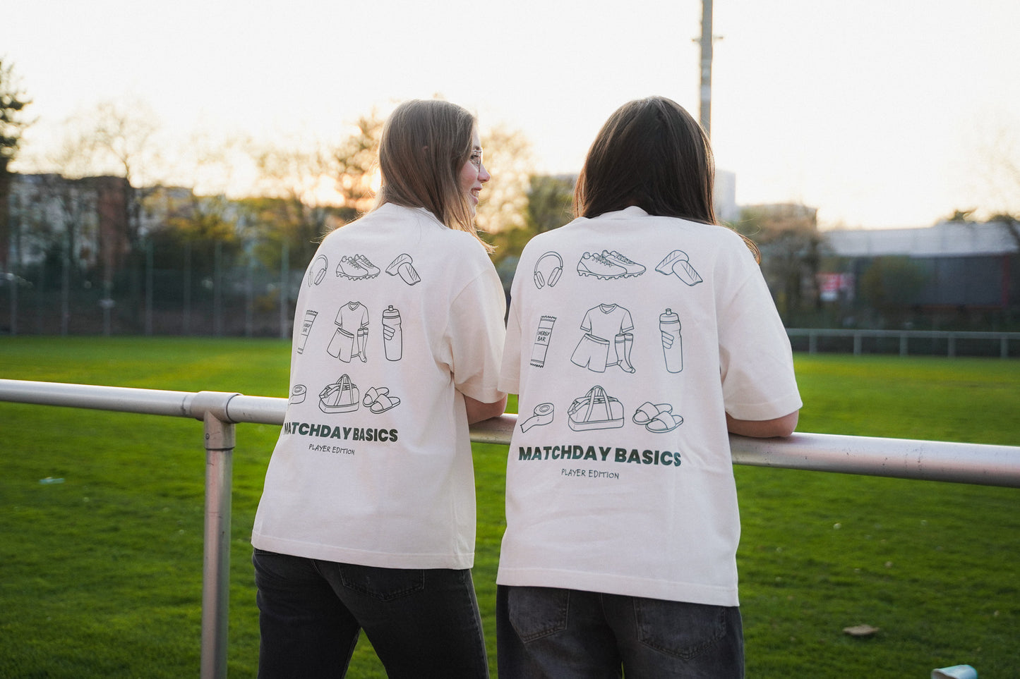 Zwei junge Frauen stehen aam Spielfeldrand eines Fußballplatzes. Sie tragen das "matchday basics" Shirt aus Chapter Three in cream.
