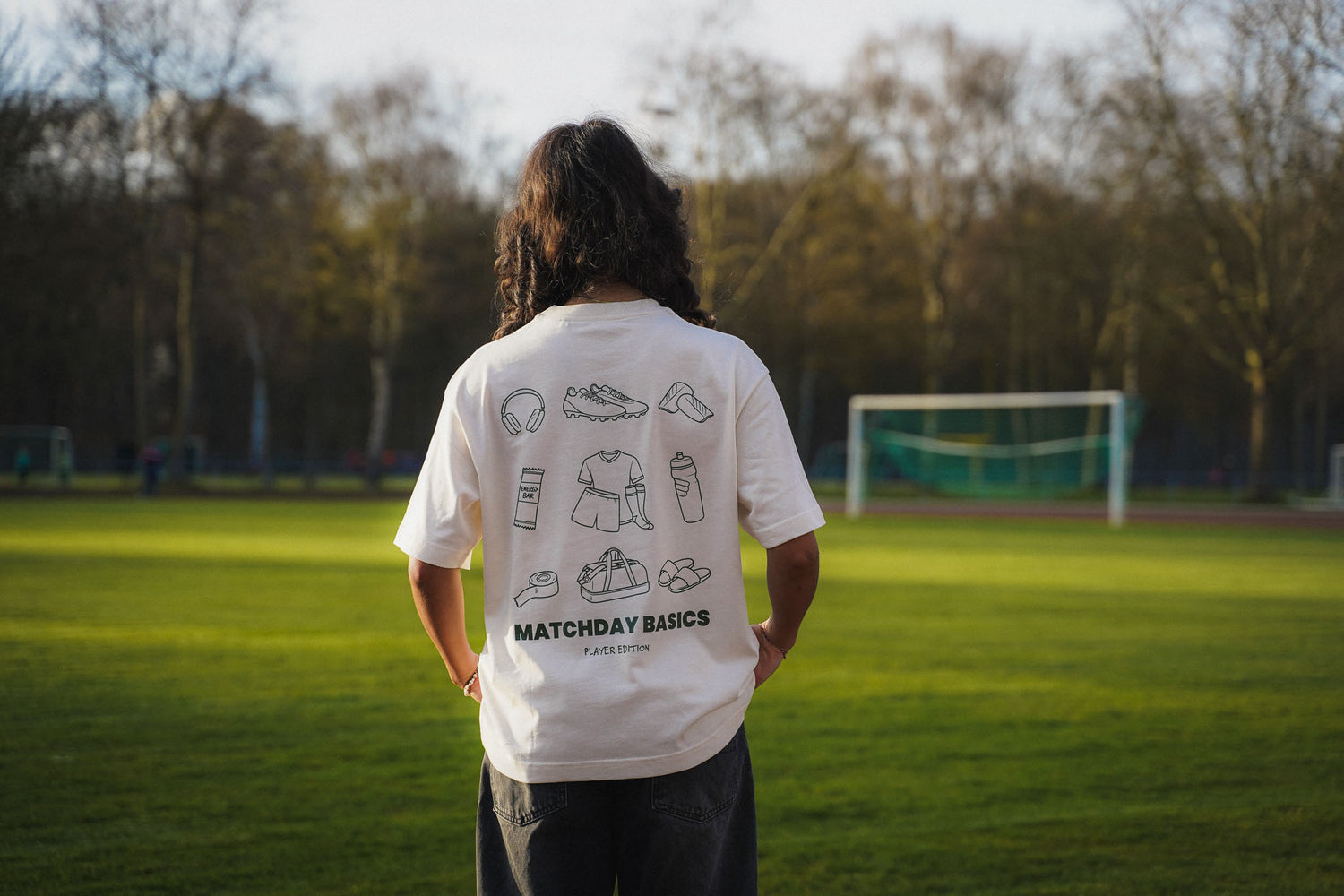 Eine junge Frau steht am Spielfeldrand eines Fußballplatzes. Sie trägt das cream Shirt mit dem Backprint "matchday basics  player edition" aus chapter three.