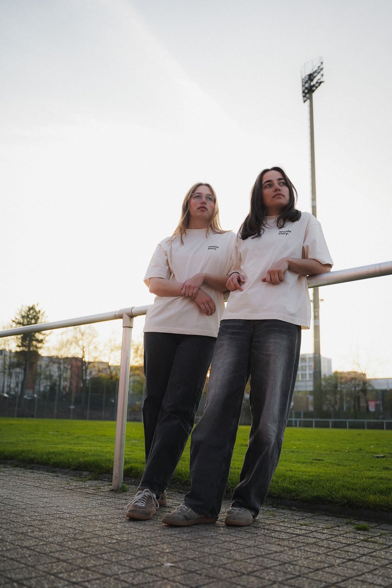 Zwei junge Frauen stehen aam Spielfeldrand eines Fußballplatzes. Sie tragen das "matchday basics" Shirt aus Chapter Three in cream.