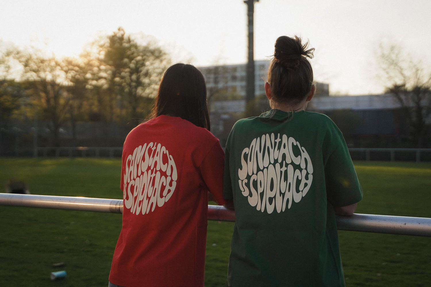 Zwei junge Frauen stehen am Spielfeldrand eines Fußballplatzes. Sie schauen in den Sonnenuntergang und tragen die Spieltach Shirts aus Chapter Three.