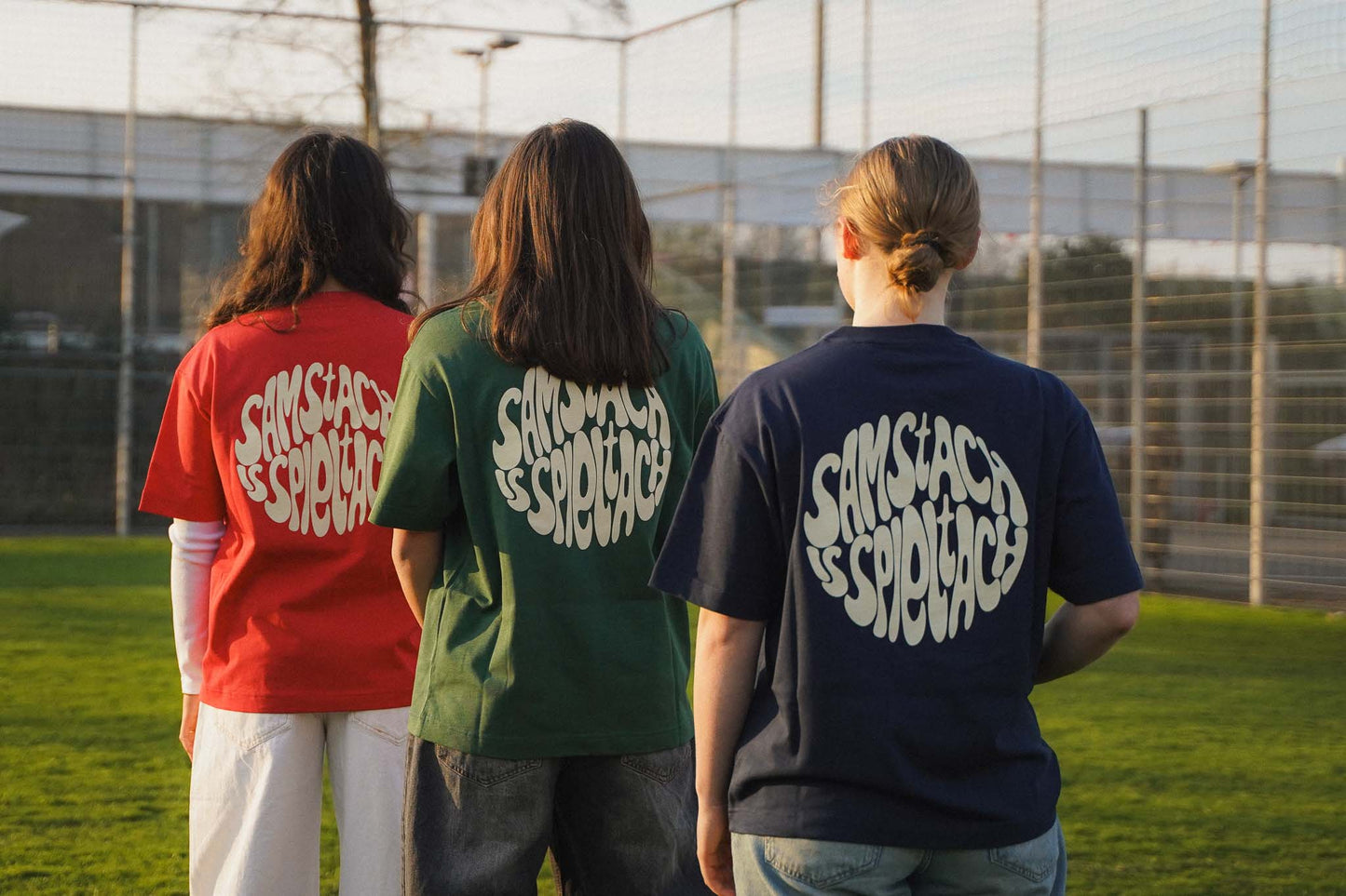 Drei junge Frauen stehen mit dem Rücken zur Kamera aufgereiht. Sie tragen alle drei Farben der "samstach is spieltach" Shirts aus Chapter Three.