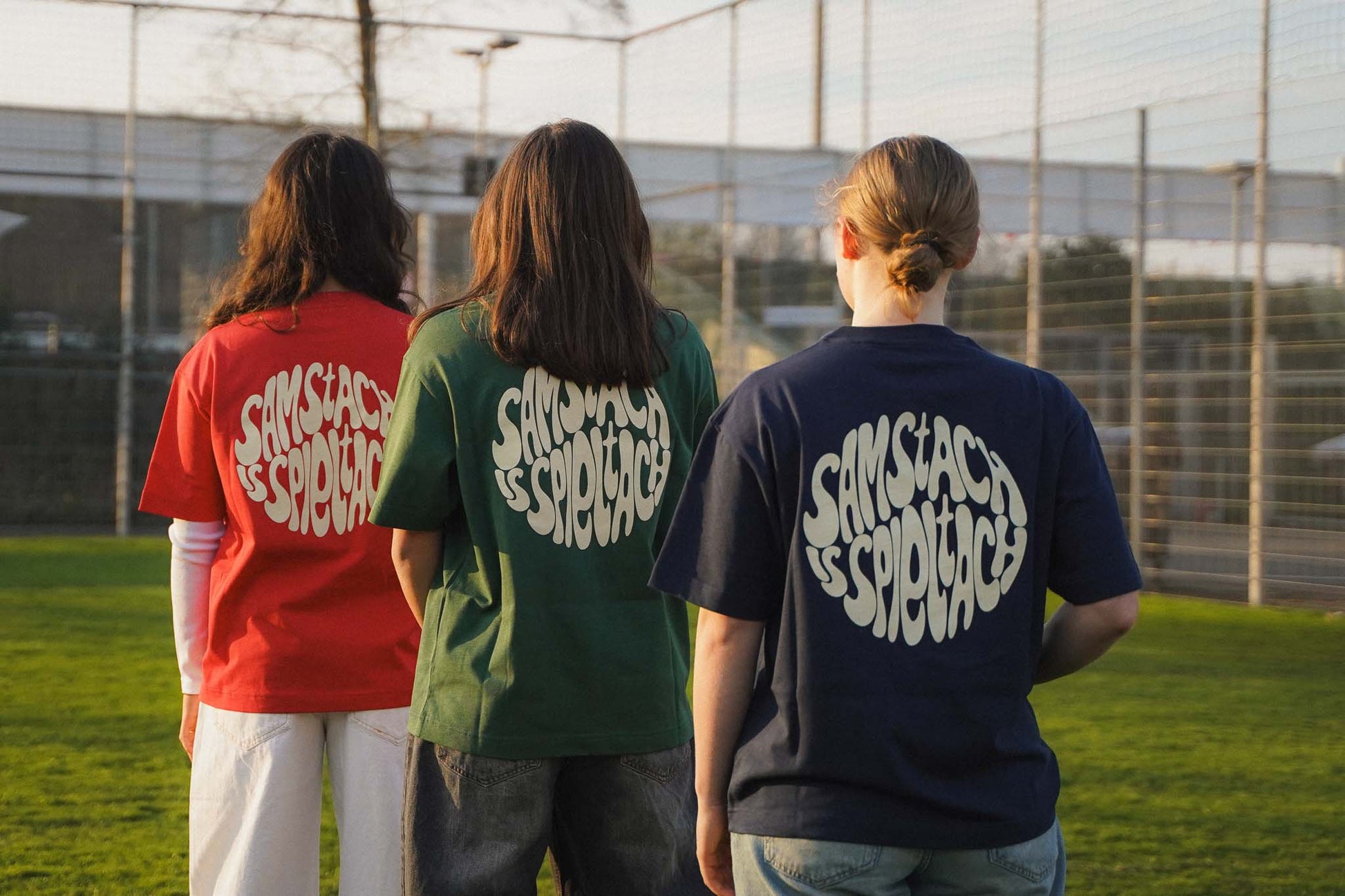 Drei junge Frauen stehen mit dem Rücken zur Kamera aufgereiht. Sie tragen alle drei Farben der "samstach is spieltach" Shirts aus Chapter Three.
