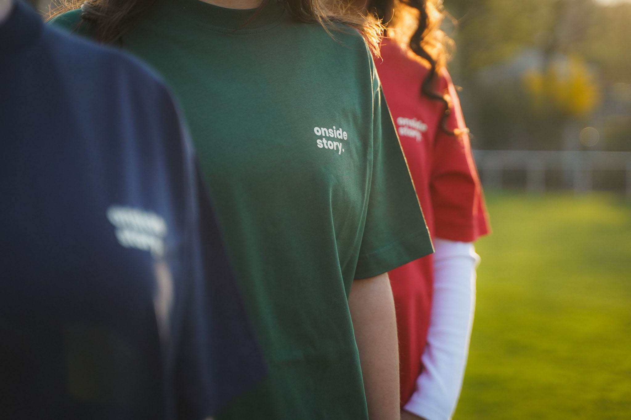 Drei Frauen stehen leicht versetzt hintereinander. Sie tragen die Spieltach Shirts in den Farben Bottlegreen, Derby Red und Midnight Blue, so dass man den Frontprint gut erkennen kann.