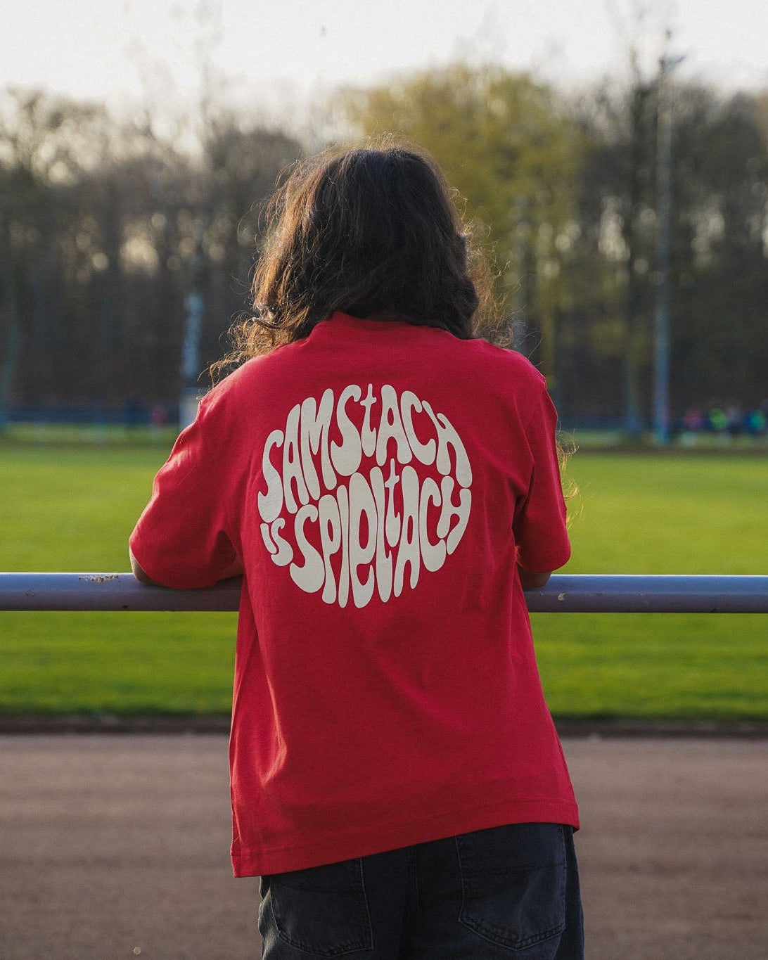 Eine junge Frauen steht am Spielfeldrand eines Fußballfelds. Sie trägt das "samstach is spieltach" Shirt in Derby Red, so dass man den Backprint sehen kann.