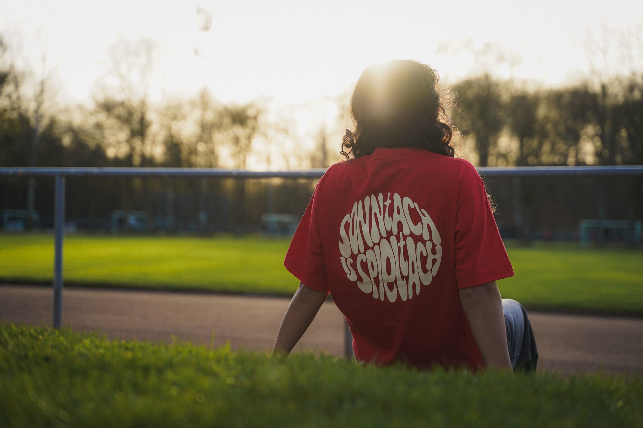 Eine junge Frau sitzt am Spielfeldrand eines Fußballplatzes und trägt das "sonntach is spieltach"-Shirt in Derby Red, so dass der Backrpint gut zu erkennen ist.