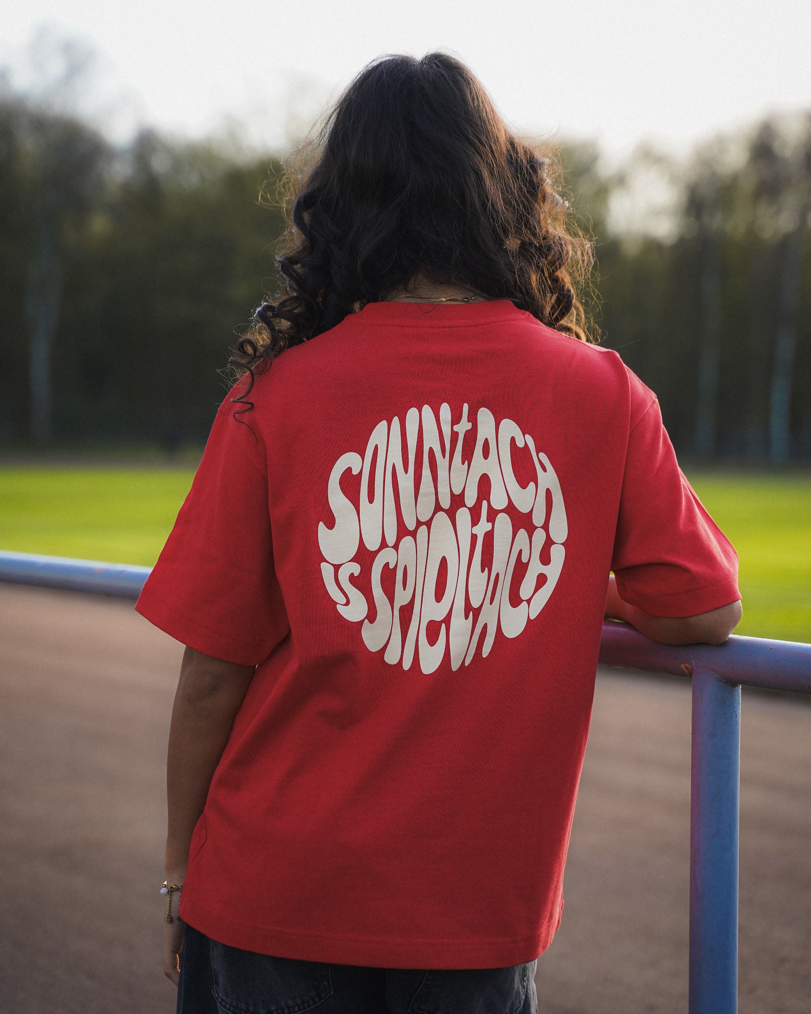 Eine junge Frauen steht am Spielfeldrand eines Fußballfelds. Sie trägt das "sonntach is spieltach" Shirt in Derby Red, so dass man den Backprint sehen kann.