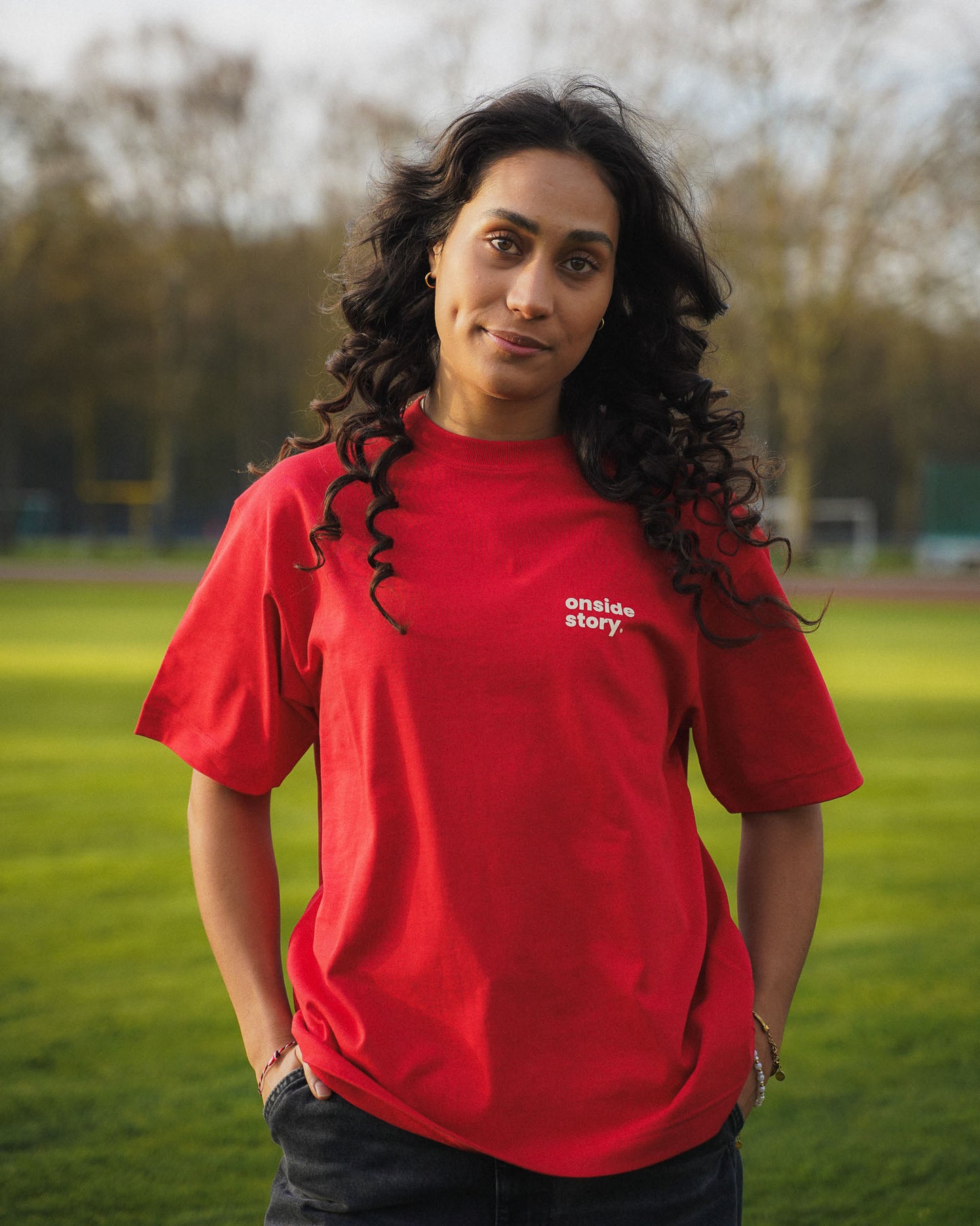 Eine junge Frauen steht auf einem  Fußballfeld. Sie schaut direkt in die Kamera und trägt das "sonntach is spieltach" Shirt in Derby Red, so dass man den Frontprint des Logos sehen kann.