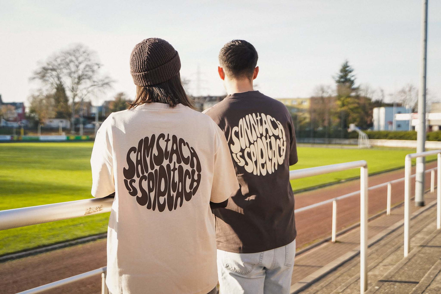 Eine junge Frau und ein junger Mann stehen am Seitenrand eines Fußballfeldes und man sieht den Backprint "samstach is spieltach" auf ihrem Shirt in Sand und "sonntach is spieltach" auf seinem Shirt in Chocolate.