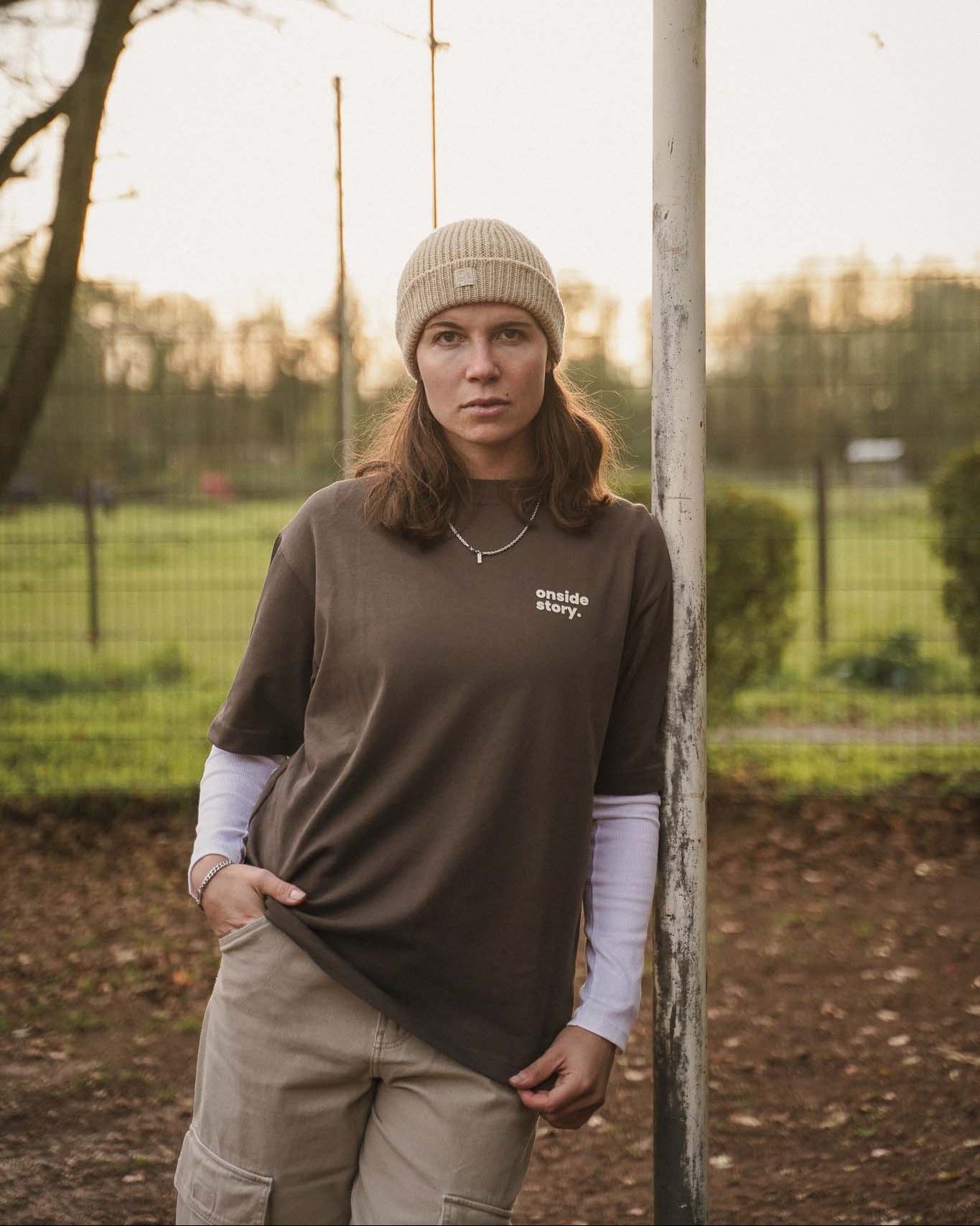 Eine junge Frauen lehnt mit der Schulter am Torpfposten eines Bolzplatzes. Sie trägt das "samstach is spieltach" Shirt in Chocolate, so dass man den Frontprint mit dem Logo gut sehen kann.