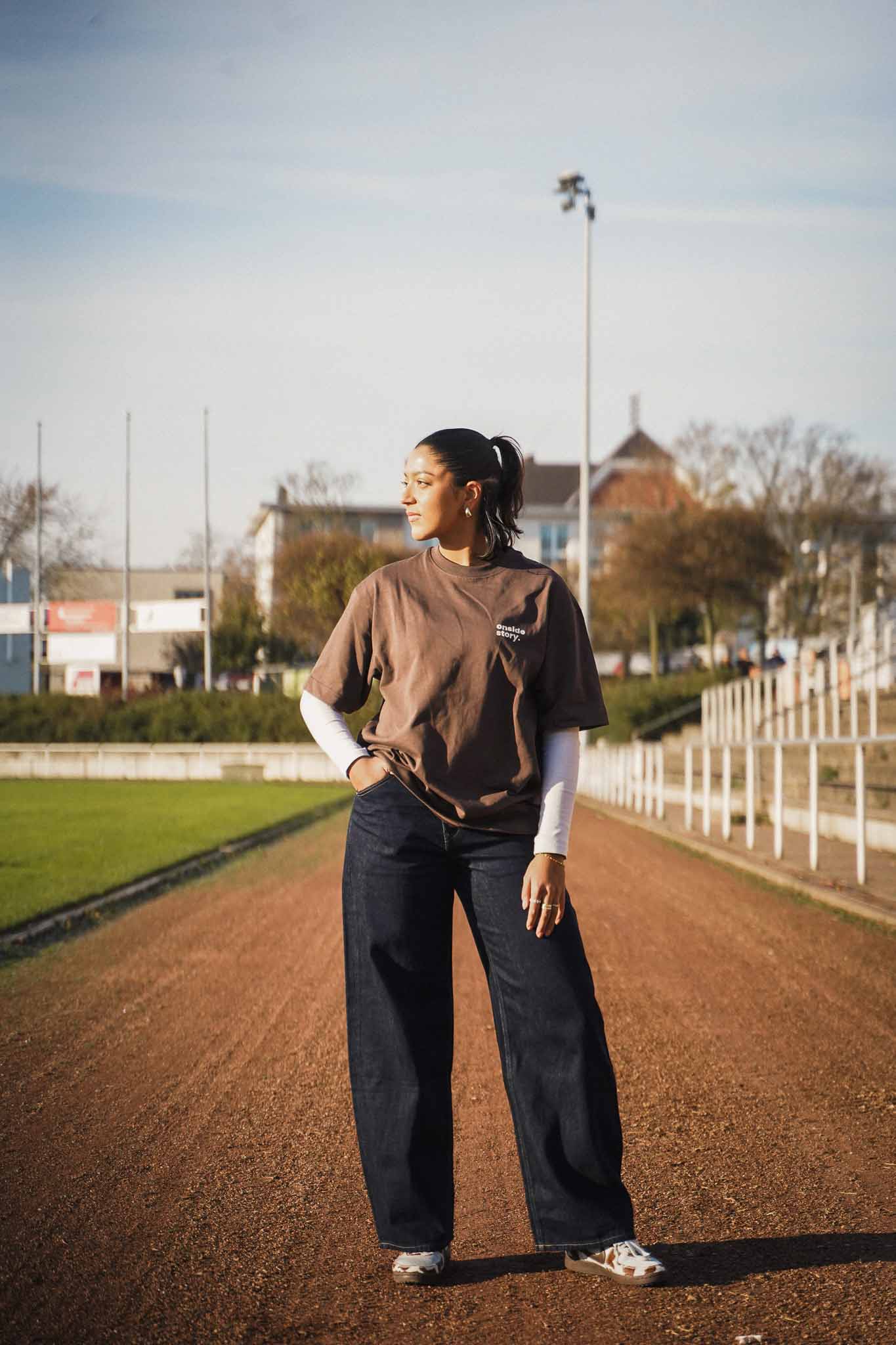 Eine junge Frauen steht auf der Asche-Laufbahn, welche ein Fußballfeld umrundet. Sie trägt das "sonntach is spieltach" Shirt in Chocolate, so dass man den Frontprint mit dem Logo gut sehen kann.