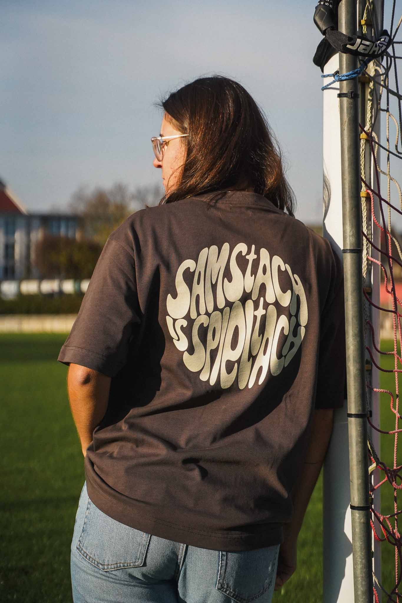 Eine junge Frau lehnt am Torpfosten eines Fußballplatzes. Sie trägt das "samstach is spieltach" Shirt in Chocolate. Der Fokus liegt auf dem Backprint.