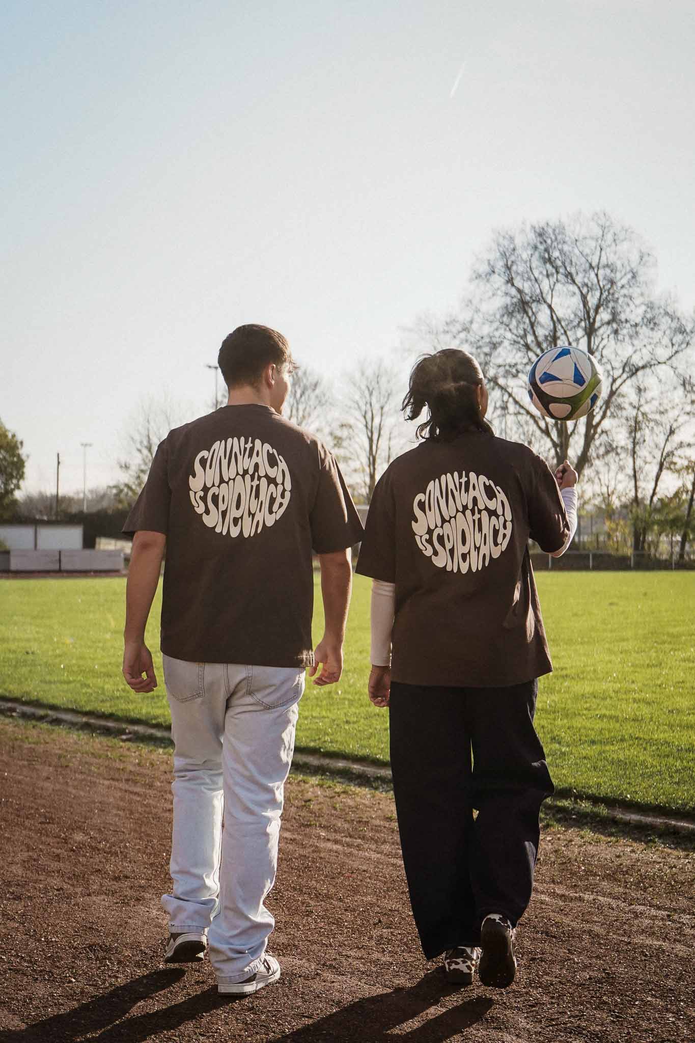 Ein junger Mann und eine junge Frau tragen das "sonntach is spieltach" Shirt in Chocolate und gehen auf ein Fußballfeld zu, so dass man den Backprint lesen kann.