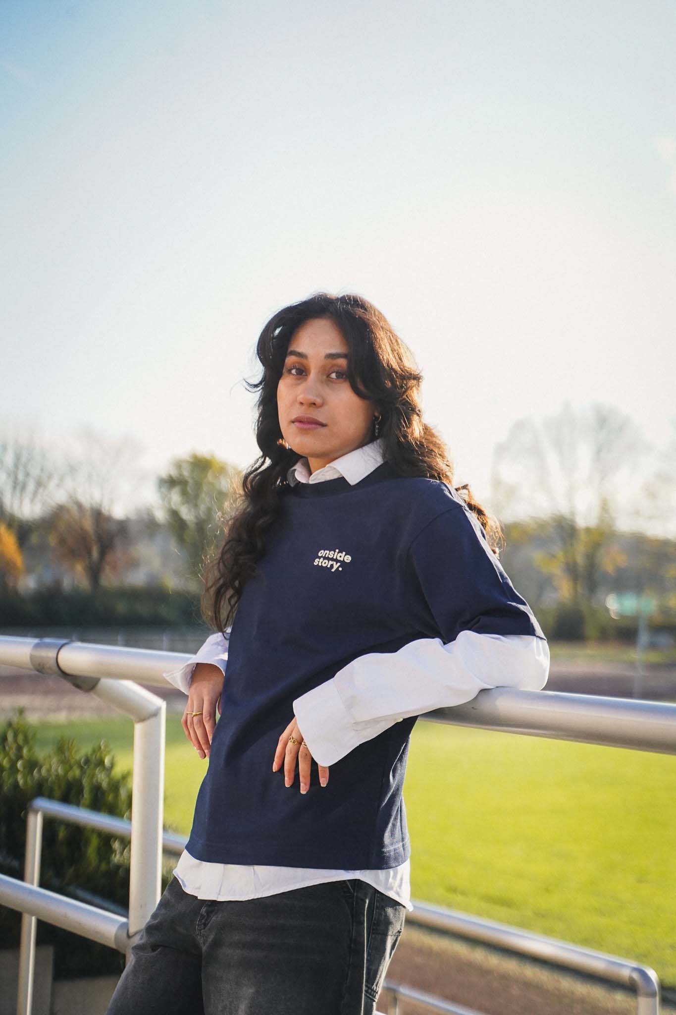 Eine junge Frauen lehnt mit dem Rücken am Stankett eines Fußballfelds. Sie trägt das "sonntach is spieltach" Shirt in Midnight Blue, so dass man den Frontprint mit dem Logo gut sehen kann.
