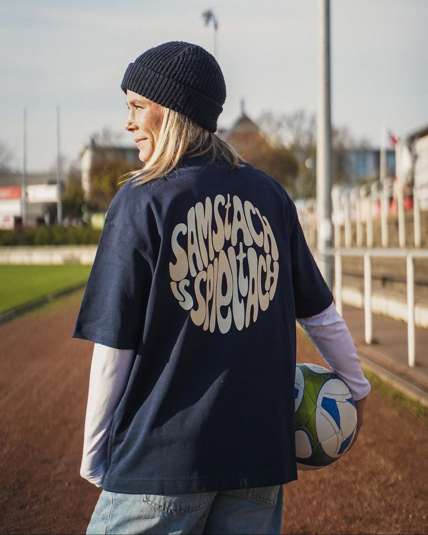 Eine junge Frauen steht auf der Asche-Laufbahn eines Fußballfelds. Sie trägt das "samstach is spieltach" Shirt in Midnight Blue, so dass man den Backprint gut sehen kann.