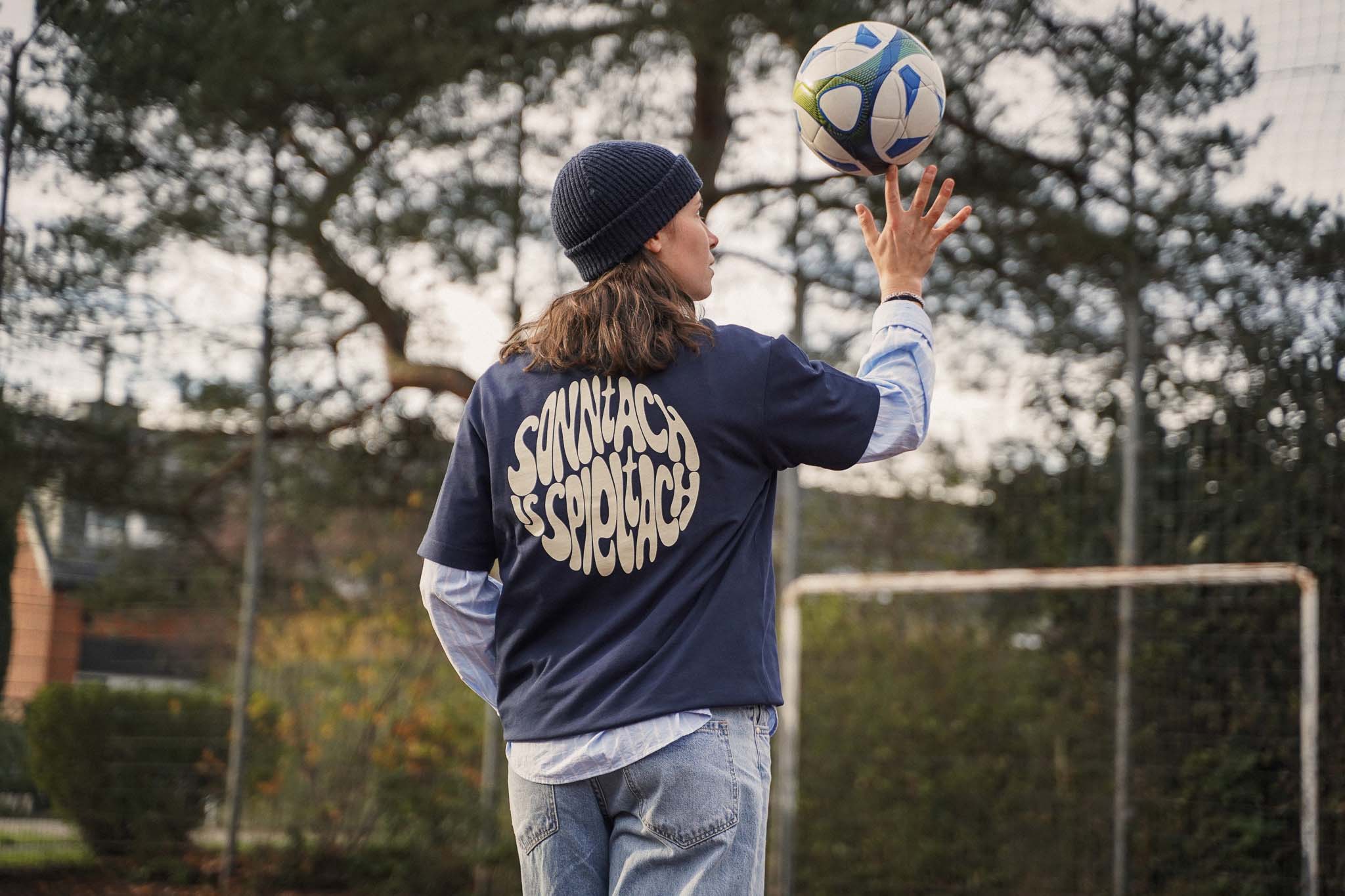 Gründerin Meret trägt das "sonntach is spieltach" Shirt in Midnight Blue auf einem alten Bolzplatz, während sie einen Fußball auf dem Finger balanciert.