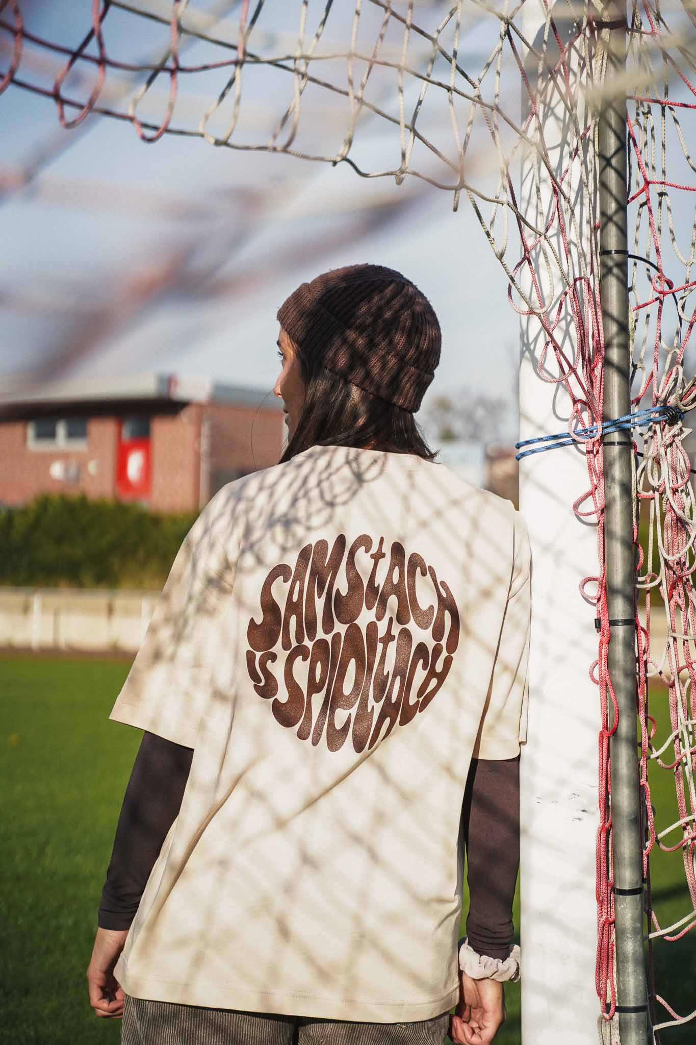 Eine junge Frauen lehnt mit der Schulter am Torpfosten eines Fußballfelds. Sie trägt das "samstach is spieltach" Shirt in Sand, so dass man den Backrpint sehen kann.