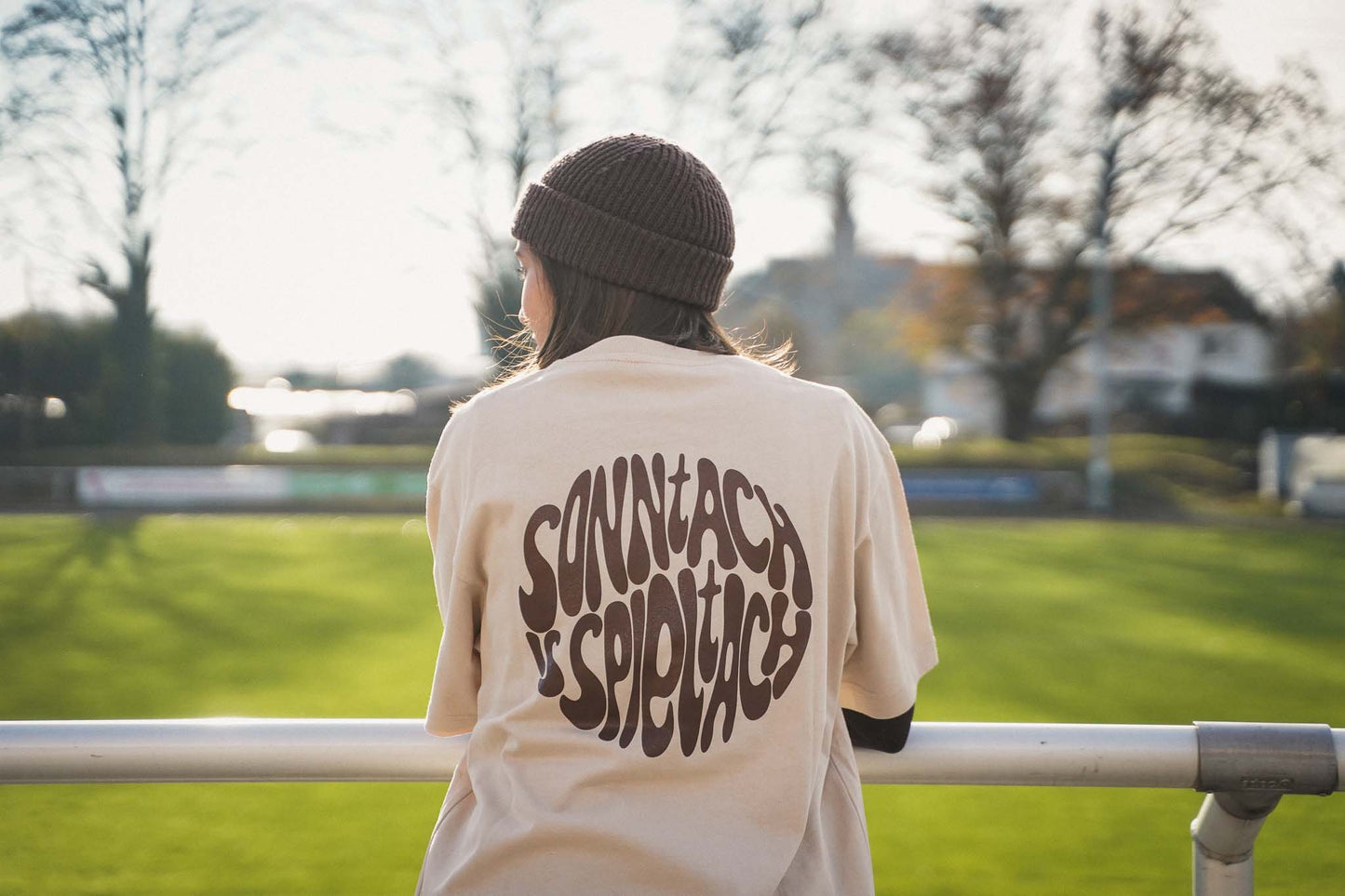 Eine junge Frauen lehnt mit den Armen am Stankett und schaut auf das Fußballfeld. Sie trägt das "sonntach is spieltach" Shirt in Sand, so dass man den Backprint in Chocolate gut sehen kann.