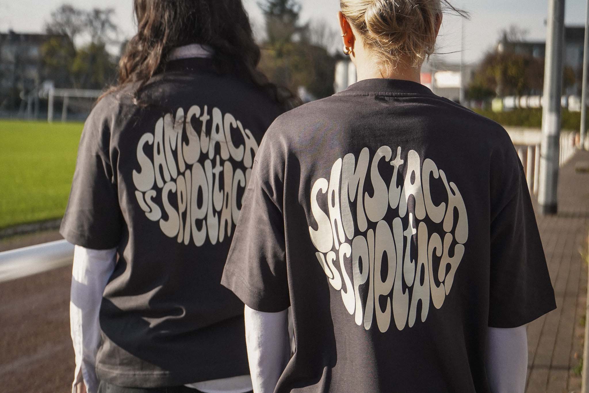 Zwei junge Frauen stehen am Spielfeldrand eines Fußballplatzes und tragen das "samstach is spieltach" Shirt in Stone Grey, so dass man den Backprint lesen kann.