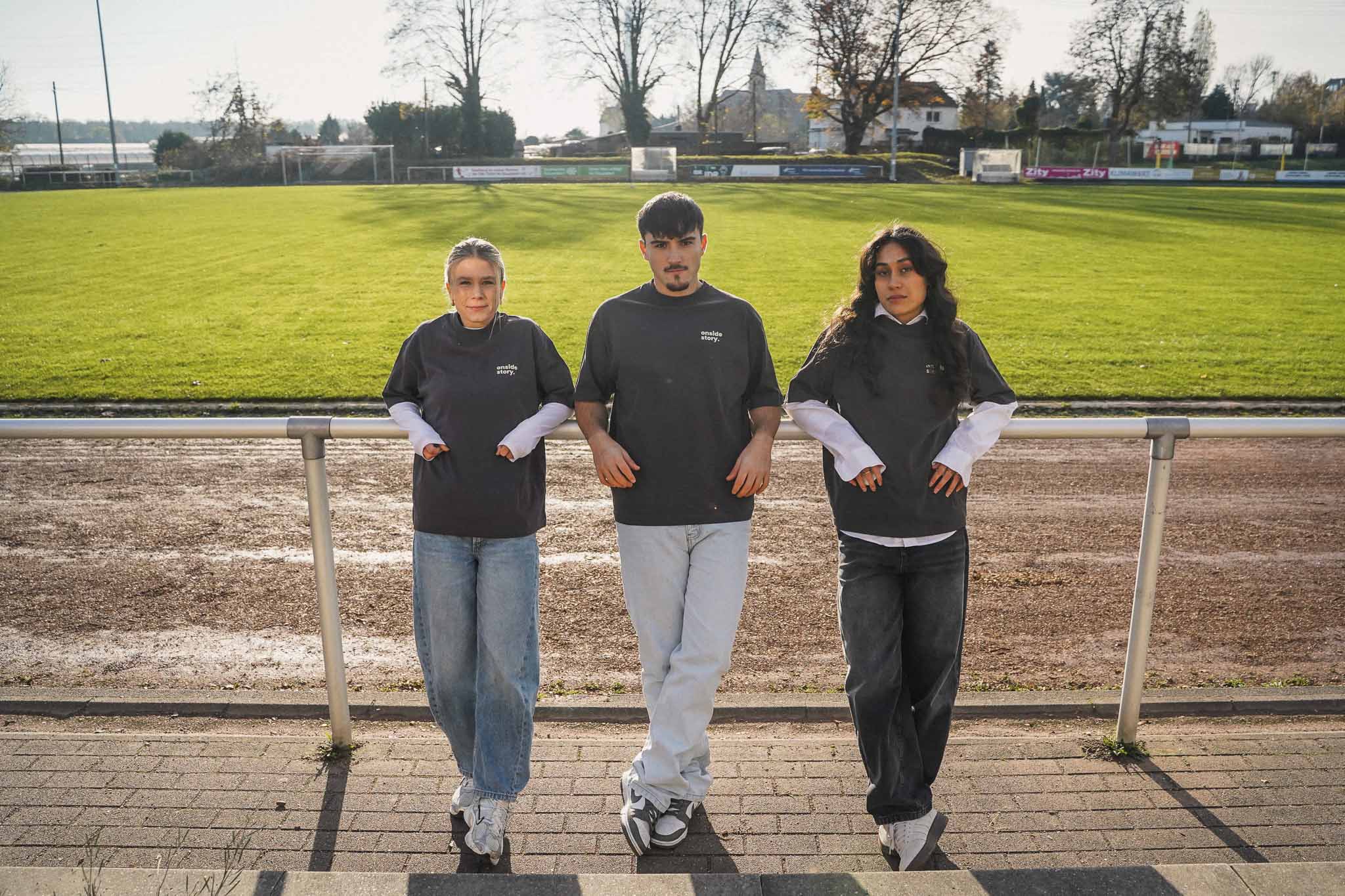 Drei junge Erwachsene lehnen mit dem Rücken am Stankett. Hinter ihnen sieht man einen Fußballplatz. Sie tragen das Shirt aus Chapter Two in Stone Grey.