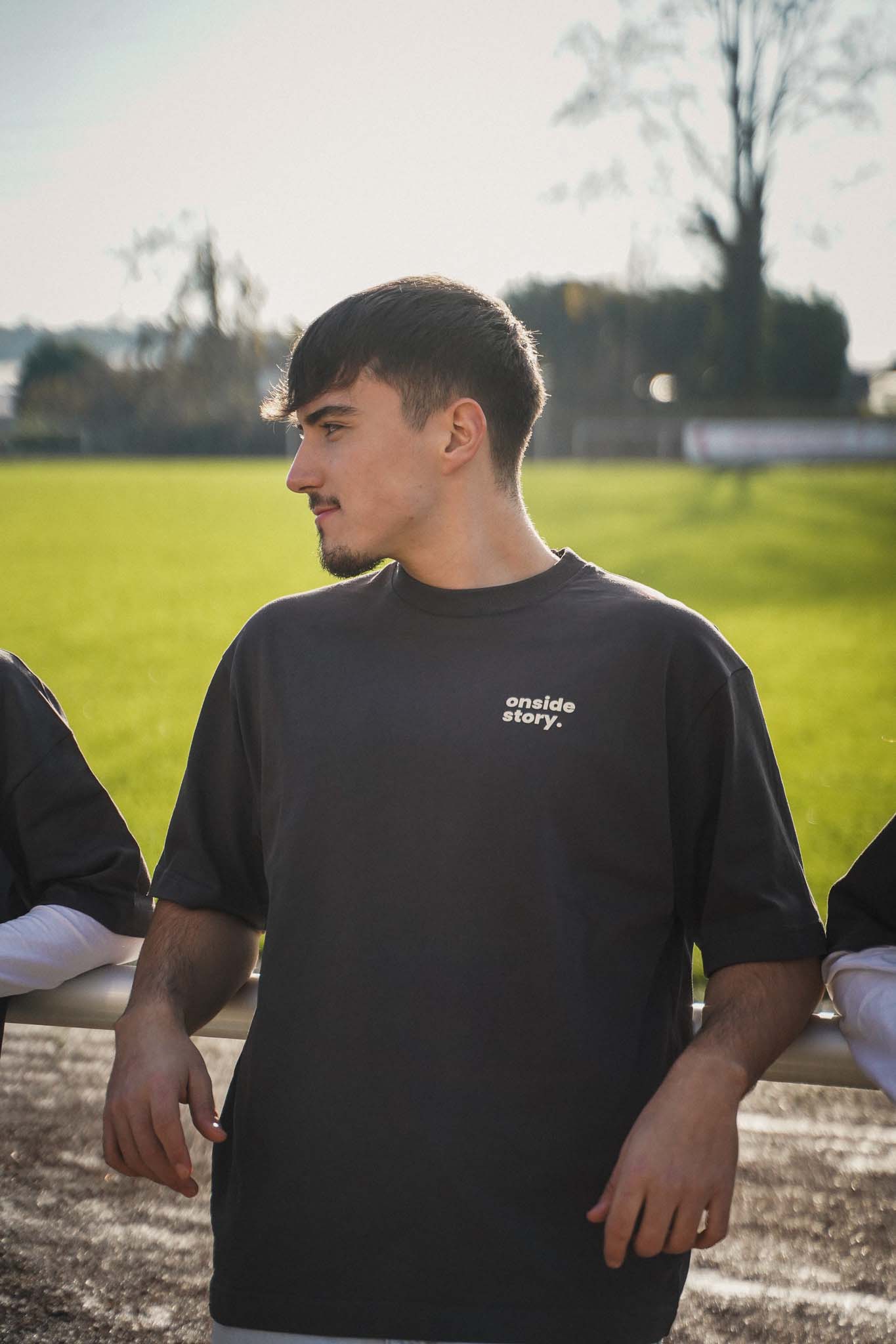 Ein junger Mann lehnt mit dem Rücken am Stankett des Fußballplatzes. Er trägt ein Shirt aus Chapter Two in Stone Grey.
