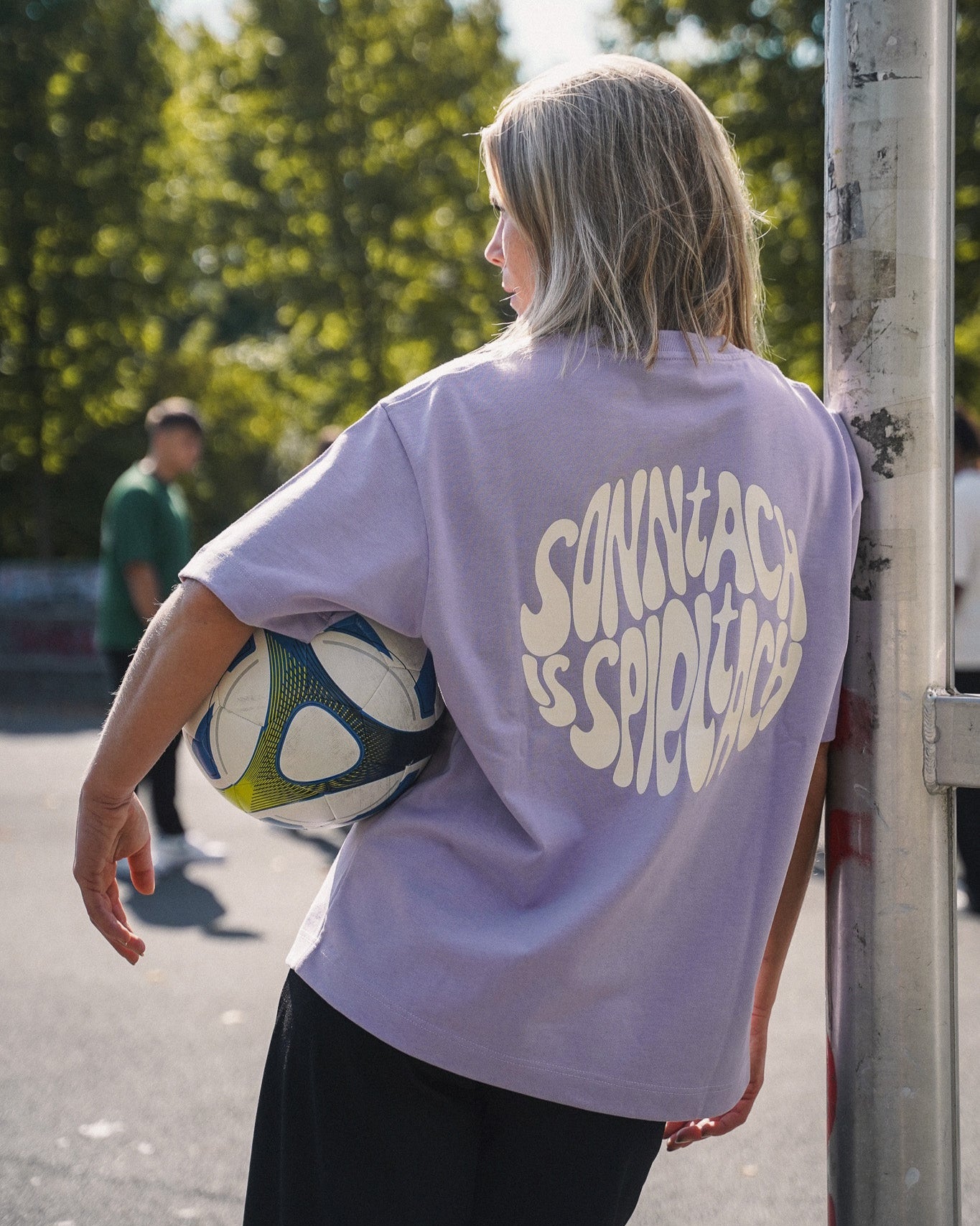 Model (160 cm) trägt nachhaltiges und fair produziertes onside story Shirt in Lavender mit Aufdruck „sonntach is spieltach“ (Größe XS) und Fußball unterm Arm.