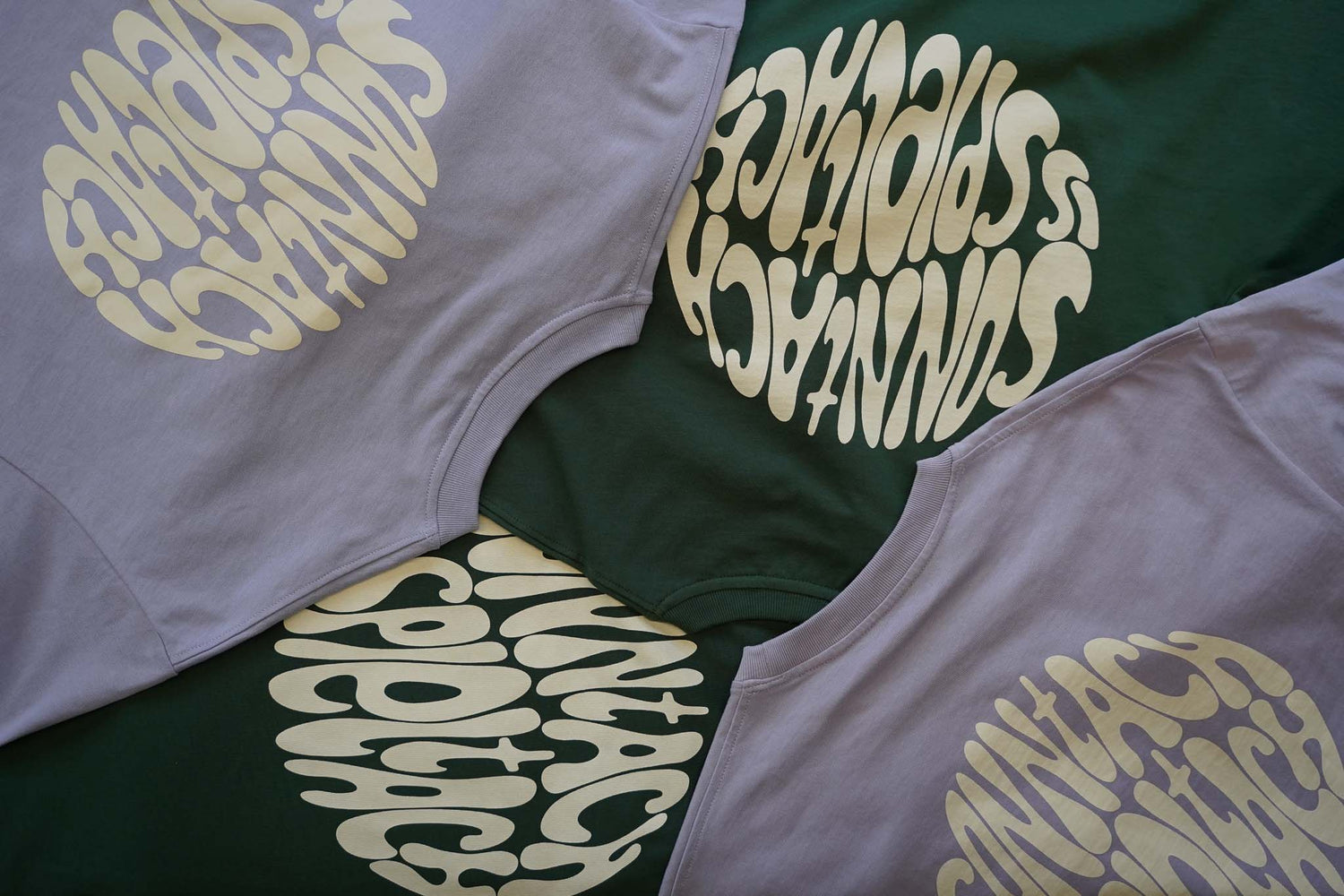 Zwei Bottlegreen und zwei Lavender Shirts liegen auf einem Tisch, so dass man den Cream Aufdruck "sonntach is spieltach" erkennt.