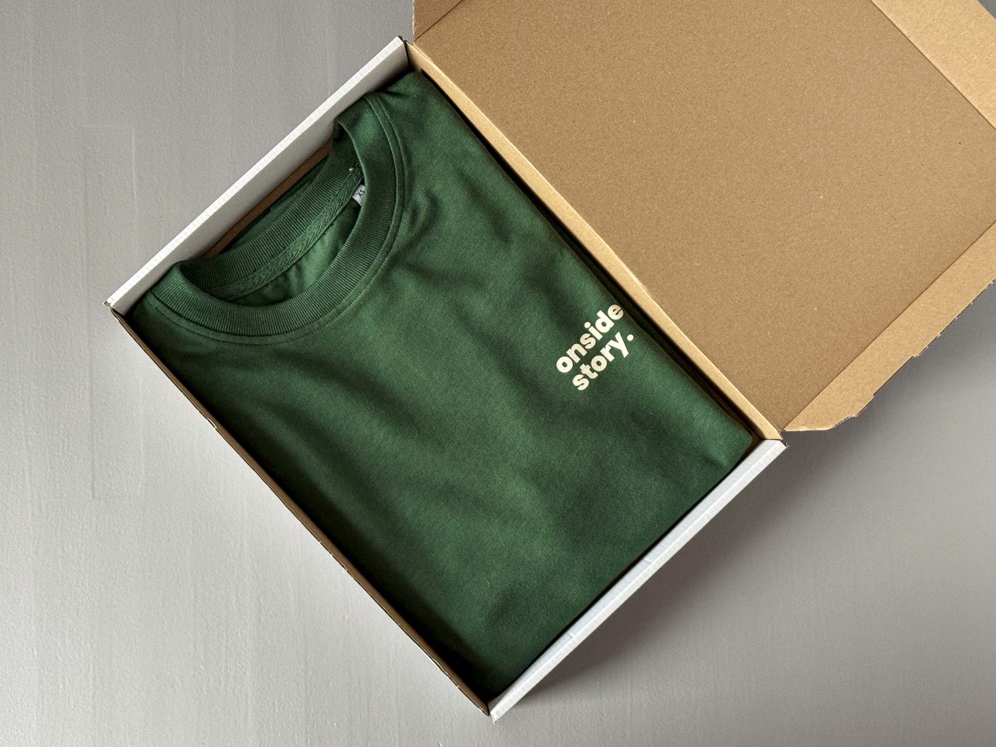 Ein Bottlegreen "sonntach is spieltach"-Shirt liegt gefalten im Versandkarton.