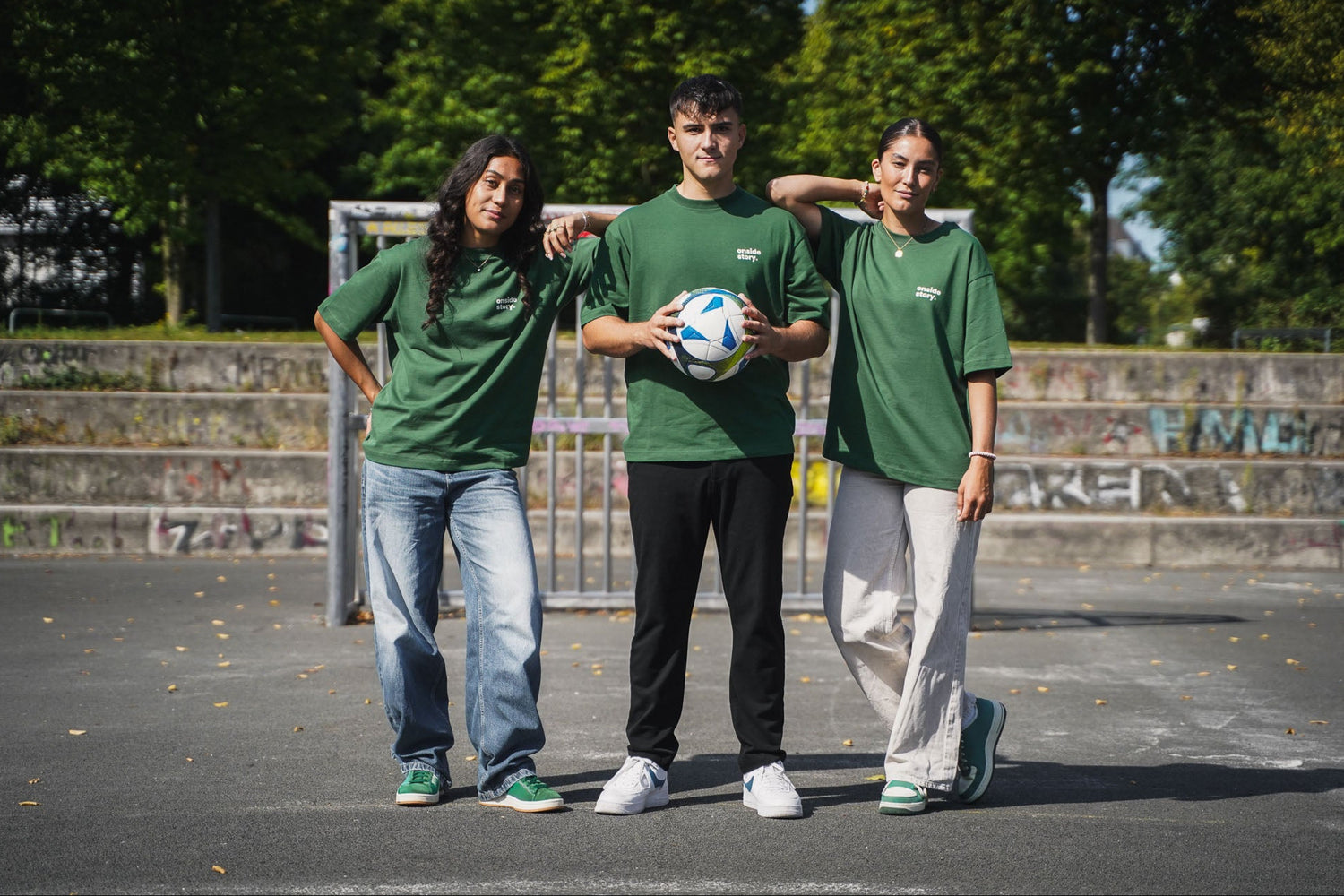 Drei Personen in den nachhaltig produzierten onside story Bottlegreen Shirts posieren mit Fußball auf einem Bolzplatz.