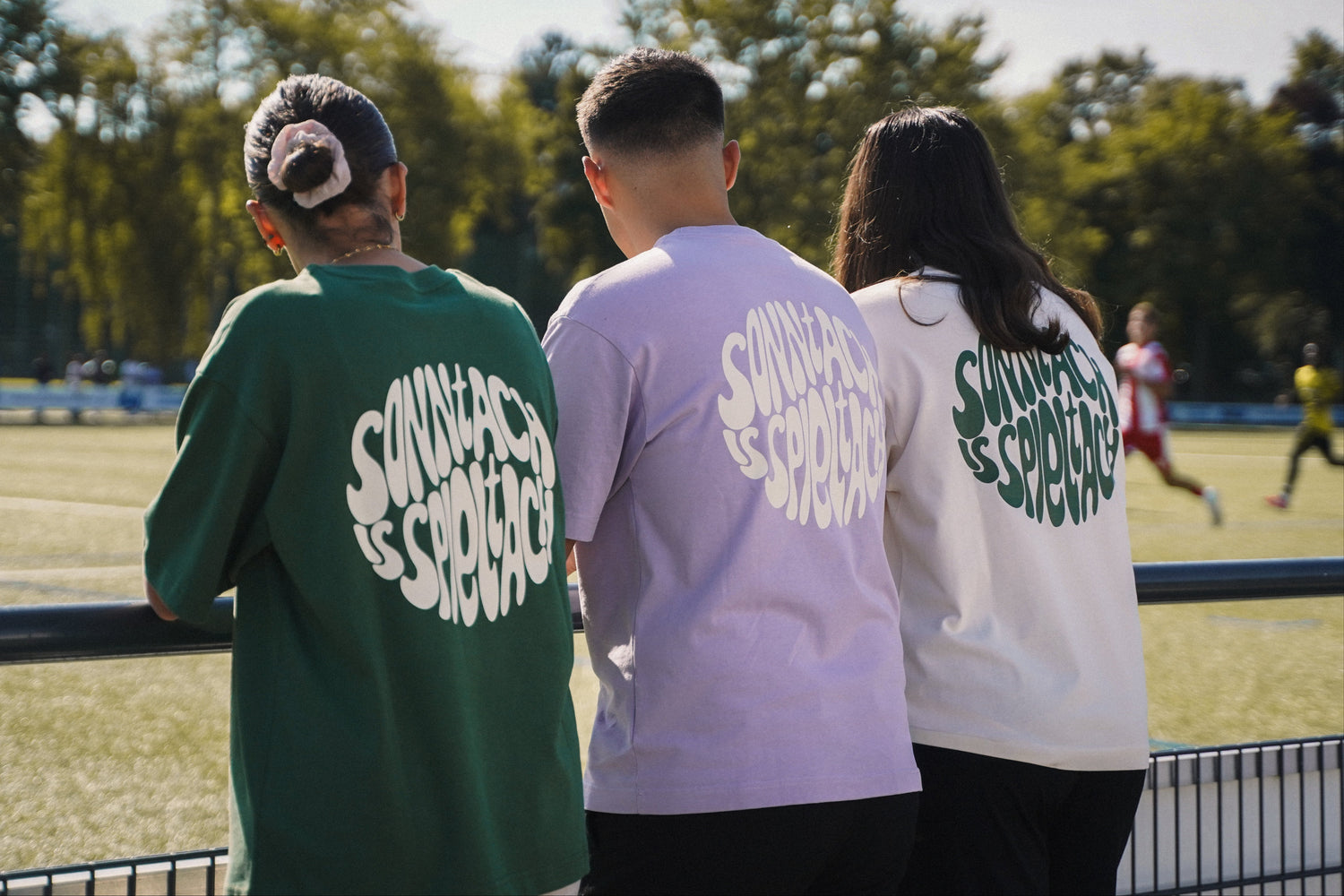 Drei Fußballfans in verschiedenfarbigen "sonntach is spieltach" Shirts schauen ein Fußballspiel.